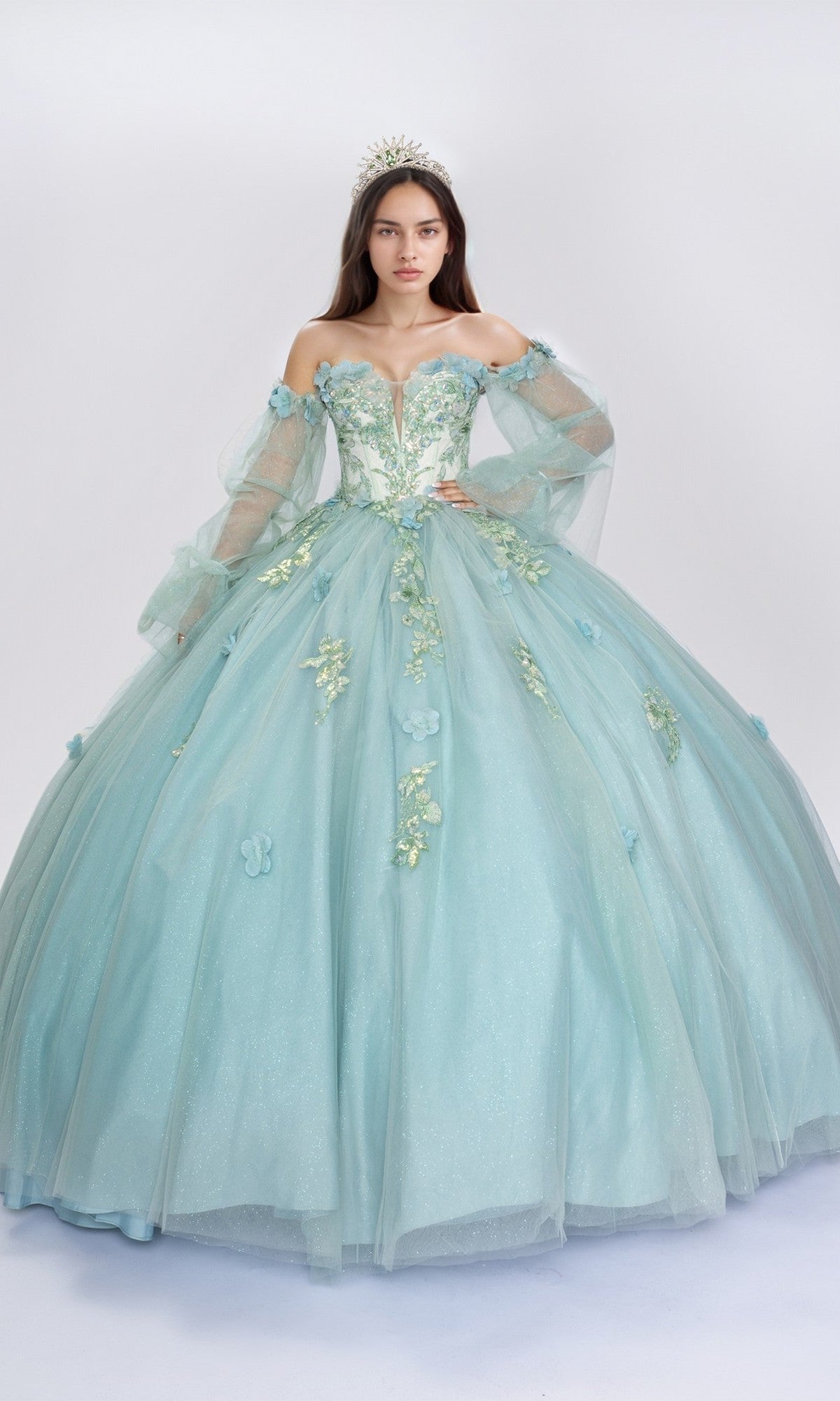 A woman in the Quinceanera Dress Q121 by Nina Canacci, a sage off-the-shoulder gown with 3D floral appliques and butterfly accents, plus a silver tiara, stands against a plain white background.